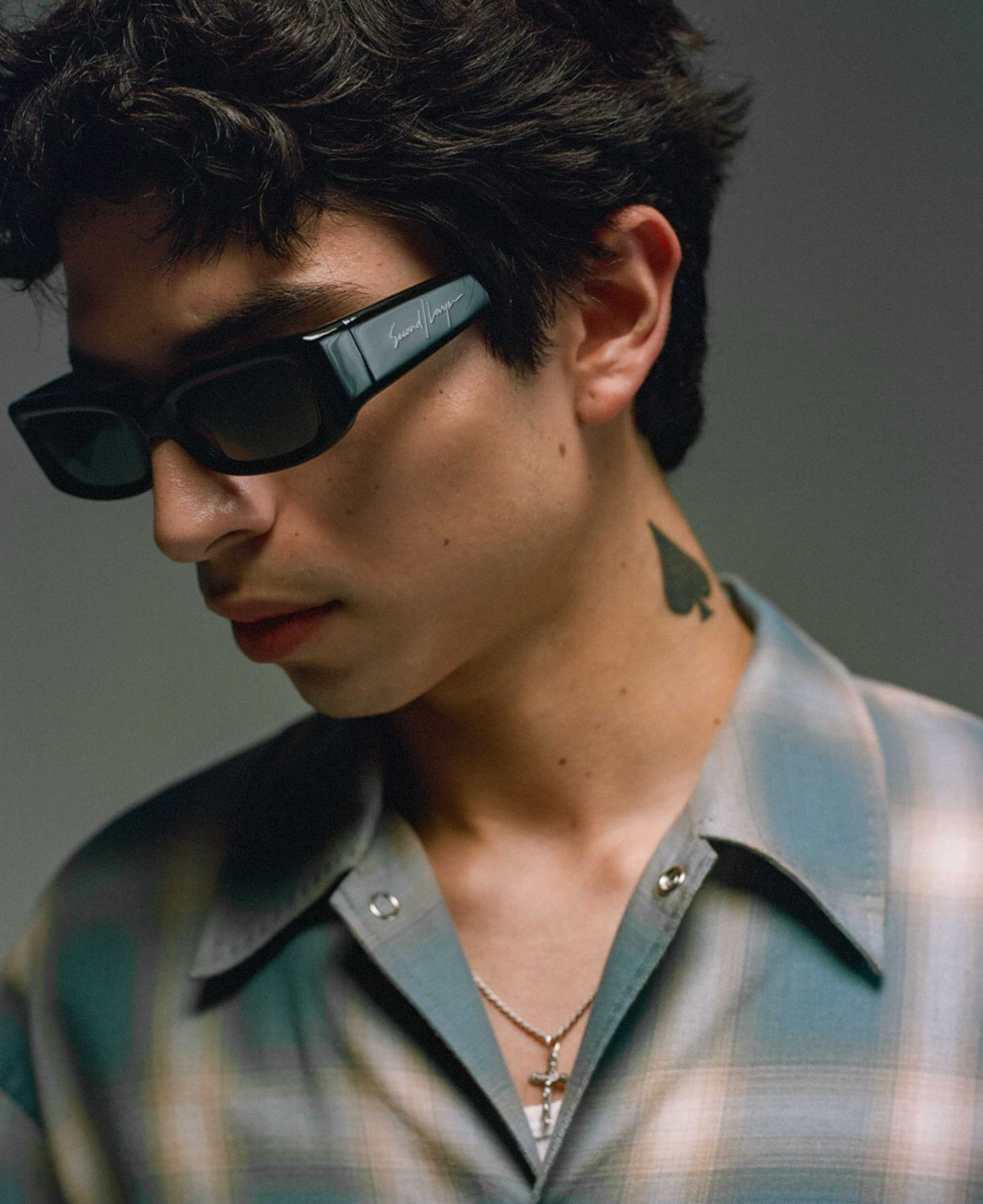 A stylish young man wearing black rectangular sunglasses with a signature detail on the side, a plaid button-up shirt, and layered silver necklaces. He has a spade tattoo on his neck and a confident, moody expression, captured in soft lighting.